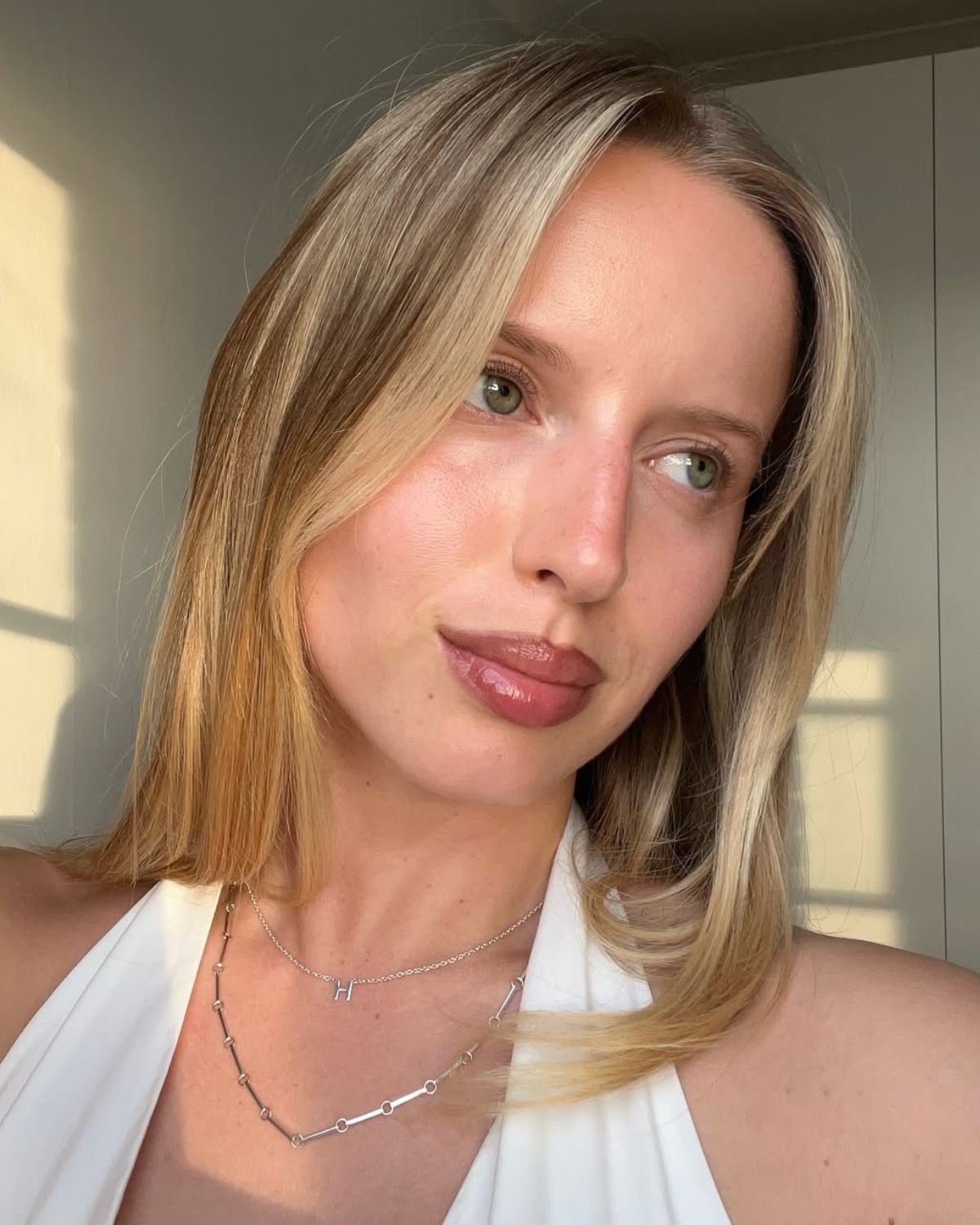 Ujęcie z bliska młodej, blond kobiety w białym topie z dekoltem typu halter, z subtelnym, świetlistym makijażem, noszącej srebrny naszyjnik z zawieszką w kształcie litery H oraz drugi, geometryczny naszyjnik; opis idealnie pasuje do artykułu o naturalnym pięknie, pielęgnacji anti-aging oraz kremy o działaniu botoksu.