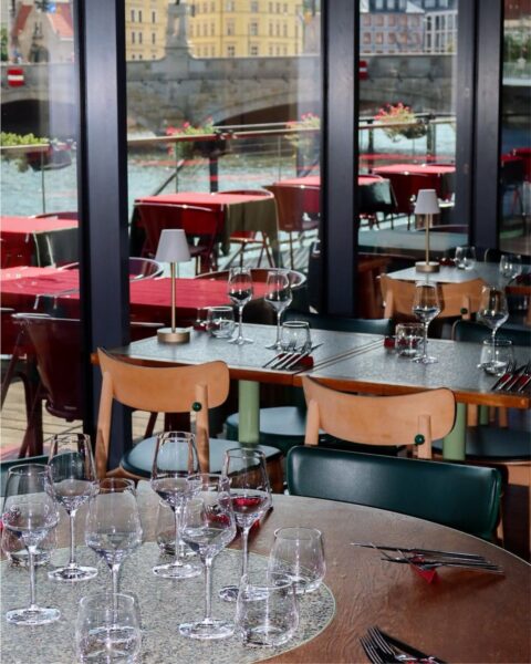 Bliski plan eleganckiego wnętrza i oszklonego tarasu, w którym znajduje się stolik z kieliszkami do wina oraz widok na rzekę i kamienną architekturę. Ta nowoczesna i stylowa restauracja Martim (Wrocław) zachwyca designem z drewnianymi krzesłami i intensywną zielenią, będąc kulinarną inspiracją dla kobiet ceniących dobry gust i atmosferę.