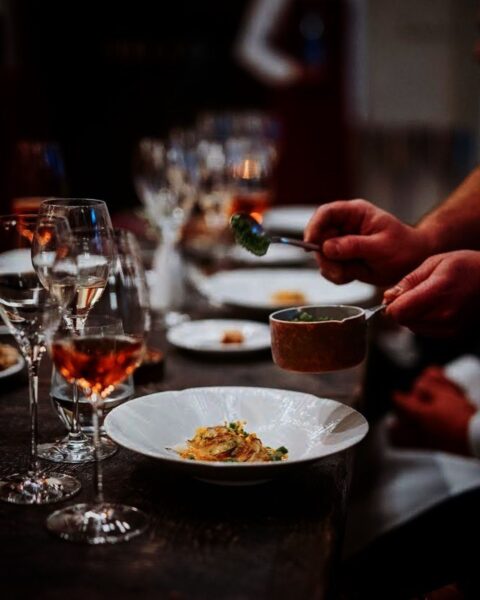 Klimatyczny, ciemny kadr z serwowania dania, podkreślający moment luksusowego doświadczenia kulinarnej celebracji. W tle widoczne kieliszki z winem, a dłonie kelnera dodają na talerz ostatnie detale. To kwintesencja wyrafinowanej atmosfery, jaką oferuje gwiazdkowa restauracja Bottiglieria 1881 (Kraków) – inspiracja dla kobiet szukających perfekcji i wyjątkowych przeżyć.