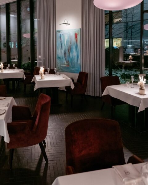 Intymne, wieczorne ujęcie wnętrza luksusowej restauracji, charakteryzujące się eleganckimi, burgundowymi, welurowymi fotelami, bielą obrusów i dużymi oknami z widokiem na miasto. Nowoczesny obraz na ścianie dodaje artystycznego sznytu. Klimat ten tworzy idealną scenerię dla wyrafinowanych kolacji, jakie serwuje gwiazdkowa restauracja Nuta (Warszawa), inspirując do celebrowania piękna w duchu emocjonalnego SEO.