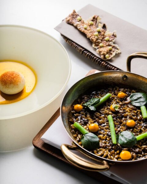 Artystycznie zaaranżowane danie fine dining, prezentujące czarny ryż w eleganckim miedzianym naczyniu, udekorowany zielonymi łodygami i żółtymi kropkami, obok jajka w kremowym sosie. Kompozycja smaku i estetyki serwowana w stylu restauracja Arco by Paco Pérez (Gdańsk) – inspiracja dla kobiet ceniących sztukę na talerzu i emocjonalną celebrację chwili.