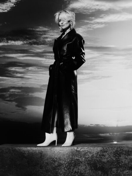 Kobieta pozuje w czarno-białym kadrze, ubrana w długi, skórzany płaszcz z paskiem i białe botki na obcasie. Na tle dramatycznego nieba prezentuje nowoczesną interpretację klasycznego stylu – siłę, elegancję i pewność siebie.