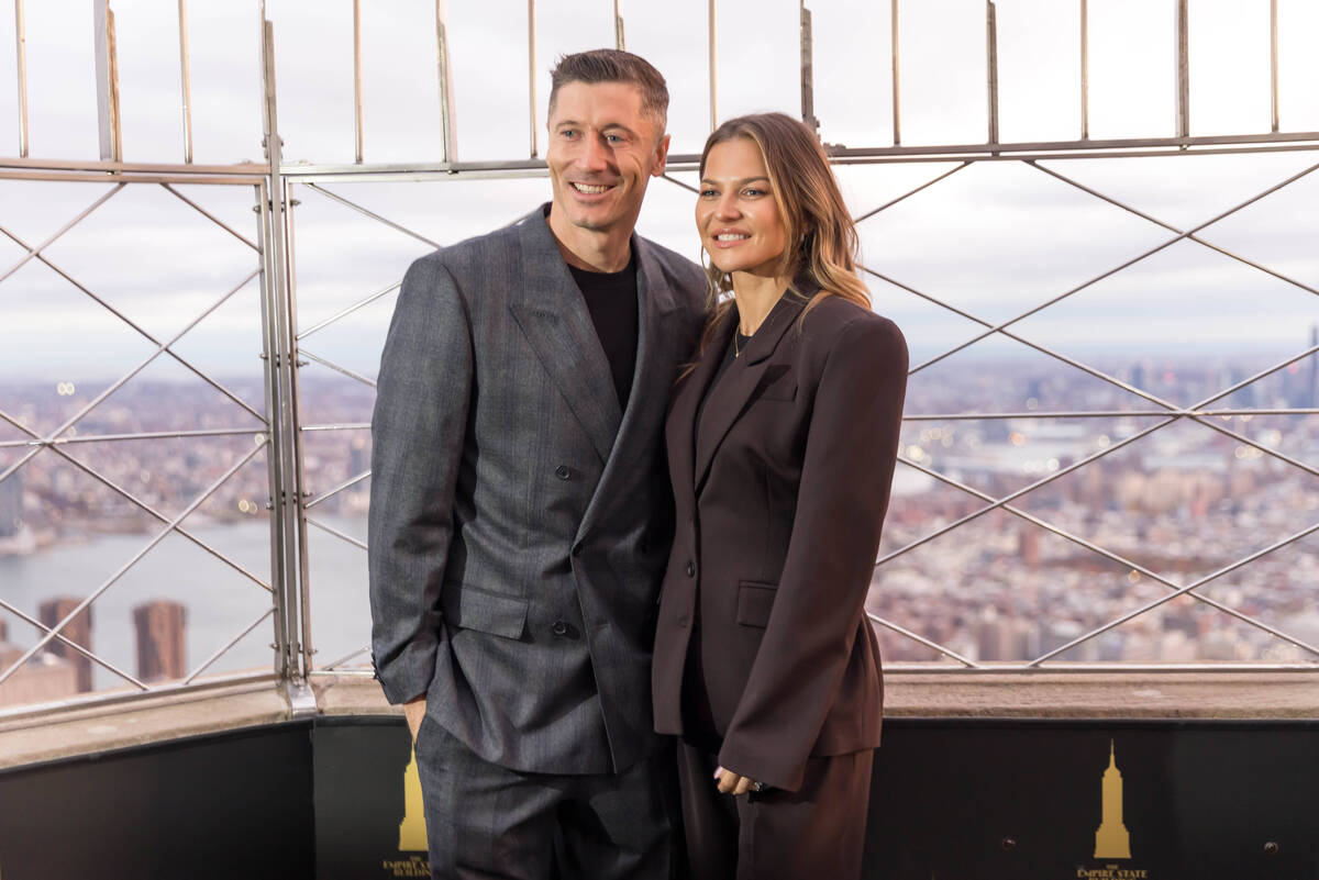 Lewandowscy na Empire State Building. Na zdjęciu widać parę, Roberta i Annę Lewandowskich na Empire State Building w Nowym Jorku. Ubrani w eleganckie, dopasowane garnitury, pozują na tarasie widokowym słynnego wieżowca. Jest to ujęcie z ceremonii, podczas której Robert Lewandowski symbolicznie podświetlił Empire State Building na biało-czerwono z okazji polskiego Święta Niepodległości. W tle rozciąga się miejska panorama Nowego Jorku pod pochmurnym niebem.