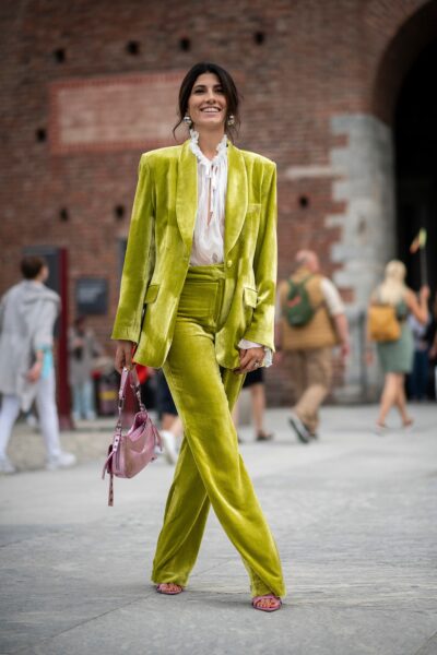 Zdjęcie przedstawia kobietę spacerującą po placu w jasnym otoczeniu miejskim, ubraną w zielony garnitur z welurową marynarką i welurowymi spodniami oraz białą bluzkę z falbanami. W tle widoczni są przechodnie oraz ceglana architektura.