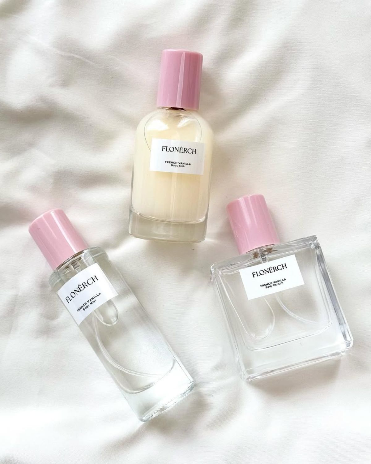 Zestaw do pielęgnacji ciała i zapachu marki FLONERCH z serii French Vanilla. Na białej, jedwabistej tkaninie leżą trzy produkty: mleczne perfumy (Body Parfum), mgiełka do ciała (Body Mist) i być może mleczko do ciała (Body Milk). Wszystkie mają jasne etykiety i różowe zakrętki. Przedstawia layering zapachowy.