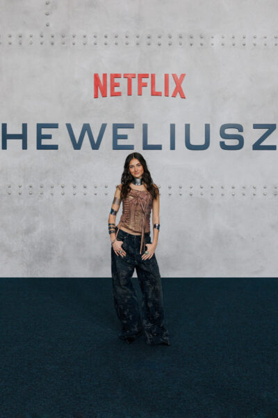 Młoda aktorka pozuje na ściance prasowej Netflixa podczas premiery serialu „Heweliusz”. Ma na sobie brązowy gorset z surowymi przeszyciami, metalowe bransolety i luźne, ciemne spodnie. Za nią widoczne logo wydarzenia na stalowym tle.