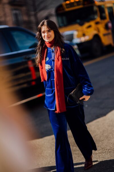 Zdjęcie przedstawia kobietę idącą ulicą w granatowej stylizacji z welurowymi spodniami, uzupełnionej czerwonym szalem i ciemnymi dodatkami. W tle znajdują się samochody oraz żółty autobus, co nadaje scenie dynamiczny, miejski charakter.