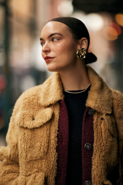 Zbliżenie na twarz kobiety w eleganckich, dużych złotych kolczykach, z gładko zaczesanymi włosami i ustami w odcieniu czerwieni; ma na sobie beżowy futrzany płaszcz i bordowy żakiet.