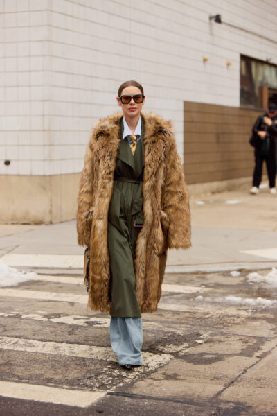 Kobieta w długim brązowo-karmelowym futrzanym płaszczu, oliwkowym kombinezonie i jasnych jeansach typu wide leg; stoi na przejściu dla pieszych, nosząc duże ciemne okulary.
