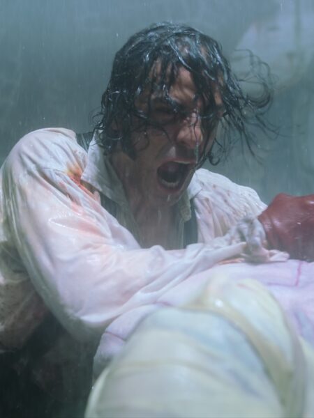 Intensywne ujęcie ukazujące mężczyznę o mokrych włosach i zakrwawionej koszuli w chwili emocjonalnego napięcia. Postać krzyczy, walcząc w deszczu, co dodaje dramatyzmu i realizmu scenie.