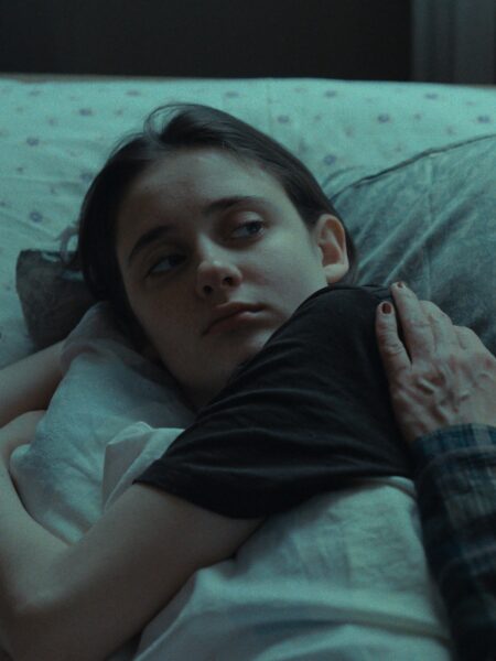 Dziewczyna leży w łóżku, oparta na poduszce, spogląda z niepokojem w bok. Kadrowanie intymne, ukazujące bliskość i delikatność relacji z osobą, której dłoń spoczywa na jej ramieniu.