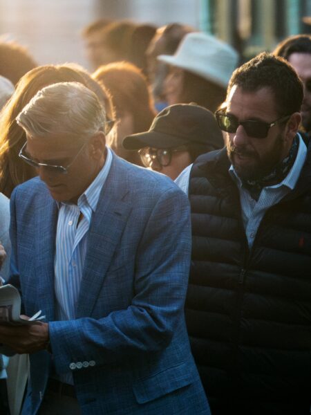 Mężczyzna w siwych włosach i okularach przeciwsłonecznych ubrany w elegancką, jasnoniebieską marynarkę w kratę oraz koszulę w delikatne paski stoi w tłumie przechodniów na ulicy o zachodzie słońca; obok niego drugi mężczyzna w czarnych okularach, pikowanej kurtce i apaszce, w tle rozmyci ludzie i ciepłe światło, tworzące atmosferę miejskiego wieczoru.