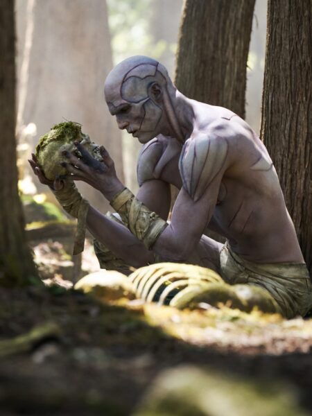 Postać o bladej, niemal szarej skórze i rzeźbiarskiej sylwetce, siedząca w lesie, trzyma w dłoniach czaszkę pokrytą mchem. Scena rozgrywa się w naturalnym świetle, wśród drzew, co potęguje atmosferę refleksji i niepokoju.