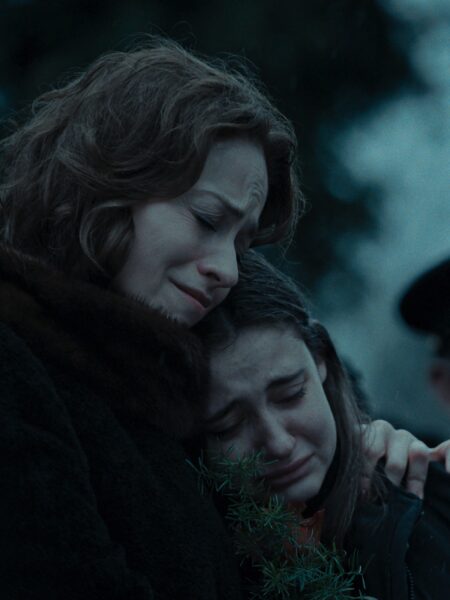 Ujęcie filmowe: kobieta przytula zapłakaną dziewczynę podczas pogrzebu, obie ubrane w ciemne, zimowe płaszcze. Scena utrzymana w chłodnej tonacji, pełna emocji i empatii.