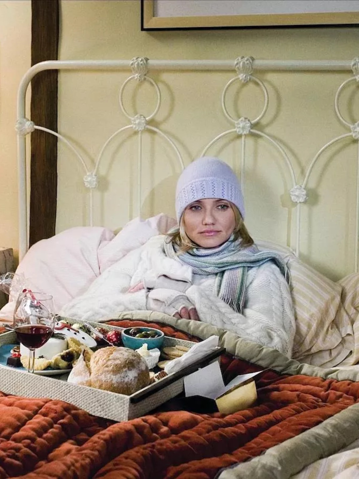 Netflix grudzień 2025: Kobieta w pastelowej czapce zimowej, grubym swetrze i szaliku siedzi w łóżku pod kilkoma warstwami koców i pościeli w ciepłych odcieniach, przed sobą ma tacę ze śniadaniem: pieczywo, sery, konfitury, kieliszek wina oraz małe miseczki z dodatkami; wnętrze sypialni jest przytulne i kojarzy się z zimowym odpoczynkiem.