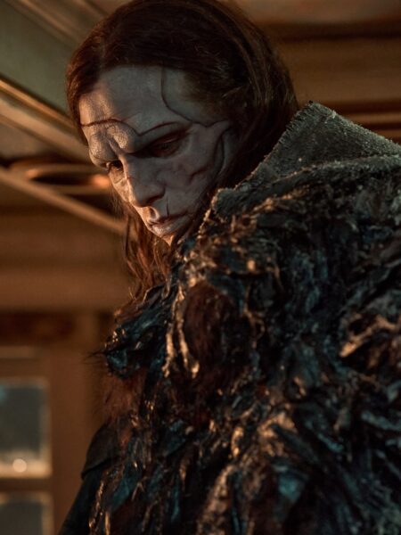 Ujęcie z półprofilu przedstawiające postać o długich włosach i zdeformowanej, blado-szarej twarzy, odzianą w ciemny, fakturowany płaszcz. Światło z wnętrza pomieszczenia tworzy kontrast, podkreślając dramatyczny nastrój.
