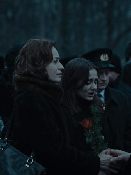 Kadr z ceremonii pogrzebowej – kobieta i dziewczyna stoją obok siebie, trzymając kwiaty i splecione dłonie. W tle widoczni uczestnicy uroczystości w czarnych płaszczach i mundurach.