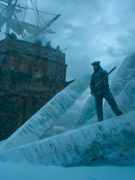 Mężczyzna ubrany w zimowy uniform stoi na skutej lodem bryle w pobliżu zamarzniętego statku o nazwie „HOPISONT”. W tle widać załogę na pokładzie i groźne, burzowe niebo. Scena utrzymana w chłodnych, niebieskich tonach.