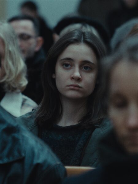 Nastolatka siedzi w kościelnej ławie, z poważnym wyrazem twarzy, otoczona tłumem dorosłych. Stonowane oświetlenie i melancholijny nastrój sceny podkreślają emocjonalny ciężar momentu.