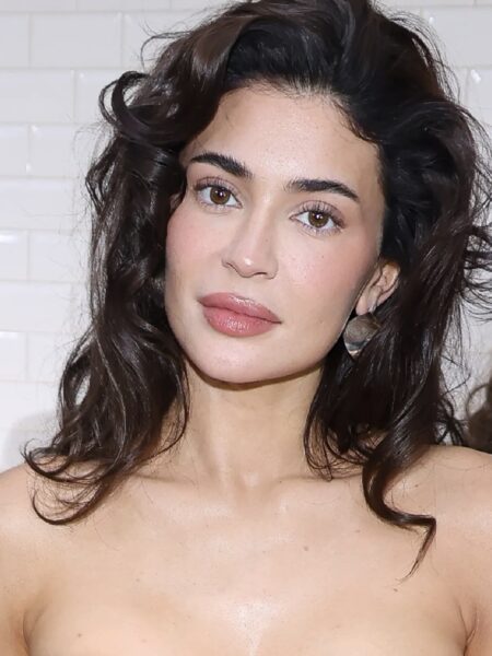 Filler fatigue co to: Kobieta w stylizacji wieczorowej – portret kobiety z ciemnymi, falującymi włosami, w delikatnym makijażu z różem na policzkach i błyszczykiem na ustach, ubranej w strapless w odcieniu nude. W tle białe kafelki i osoby w ciemnych ubraniach.