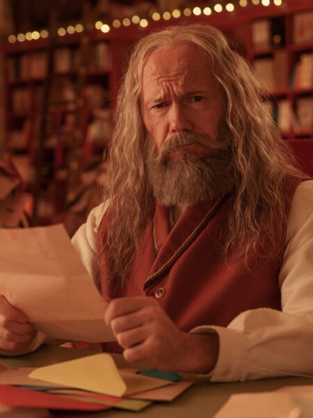 Doświadczony mężczyzna w długich siwych włosach i brodzie, ubrany w czerwony kamizelkowy strój z kremową koszulą, siedzi przy stole pełnym kolorowych kartek i listów, trzymając w dłoniach pismo i spoglądając z powagą; za nim półki z książkami oraz świąteczne światełka, tworzące ciepłą, klimatyczną scenerię.