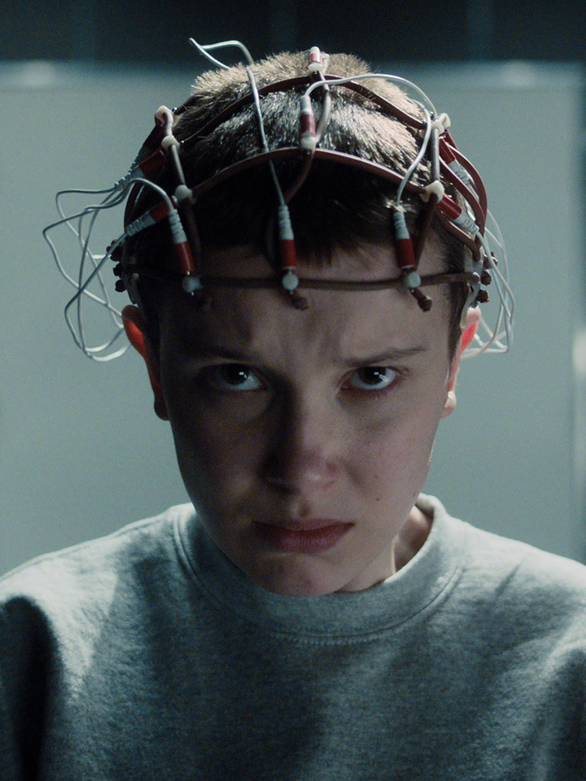 Stranger Things 5 kiedy: Kadr z serialu science fiction – młoda postać z krótkimi włosami, ubrana w szary dres, siedzi w laboratorium z surowym, chłodnym oświetleniem. Na jej głowie znajduje się metalowa opaska z przymocowanymi kablami i elektrodami, sugerująca eksperyment naukowy lub test telepatycznych zdolności. Twarz skupiona, z intensywnym, poważnym spojrzeniem w obiektyw kamery.