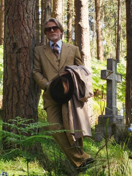 Elegancko ubrany mężczyzna w trzyczęściowym garniturze w odcieniu karmelu, z okularami przeciwsłonecznymi i kapeluszem trzymanym w dłoni, stoi oparty o pień drzewa w leśnym otoczeniu, obok starego kamiennego krzyża; miękkie światło i gęsta zieleń podkreślają melancholijny, refleksyjny nastrój kadru.