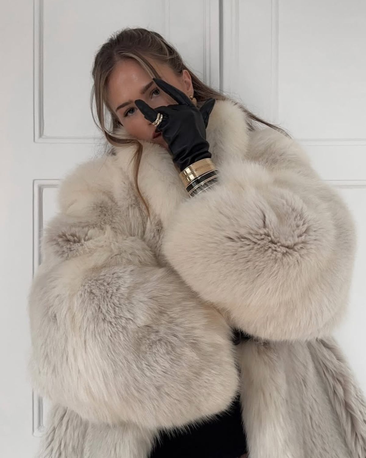 Kobieta w luksusowym, białym futrze (najprawdopodobniej z lisa lub sztucznym futrze typu faux fur) z obszernymi, bufiastymi rękawami. Ma na sobie czarne, koronkowe rajstopy i ciemne body lub strój. Na dłoni, którą zasłania część twarzy w geście pokoju, ma elegancką czarną rękawiczkę i kilka złotych bransolet. Tło stanowi prosta, biała powierzchnia (drzwi). Stylizacja jest szykowna, ekstrawagancka i ciepła. Zdjęcie idealnie pasuje do artykułu o tym, jak stylowo przetrwać chłody i dopełnić look luksusowymi detalami. To wszystko przywołuje na myśl otulające perfumy na zimę 2025.