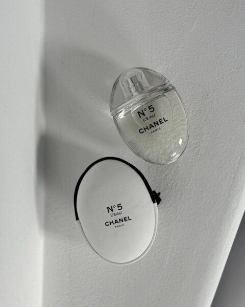 Zdjęcie przedstawia minimalistyczną kompozycję na jasnym, architektonicznym tle. Na pierwszym planie znajduje się flakon perfum Chanel N°5 L'EAU oraz pasująca do niego okrągła, biało-czarna saszetka lub etui z tym samym logo. Flakon leży obok etui na białej, zakrzywionej powierzchni, tworząc grę cieni, która podkreśla elegancję i nowoczesny minimalizm marki.