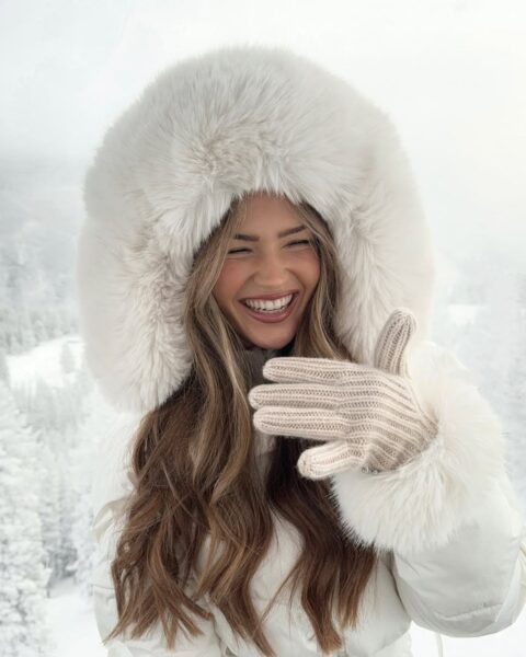 Zbliżenie twarzy (selfie) kobiety w luksusowym, białym kapturze obszytym gęstym futrem (lub sztucznym futrem). Kobieta ma długie, jasnobrązowe włosy i naturalny, promienny makijaż, skupiony na błyszczących ustach w odcieniu nude. Zdjęcie ma elegancki, zimowy charakter i idealnie ilustruje trendy w modzie narciarskiej lub pielęgnację skóry na mrozy.
