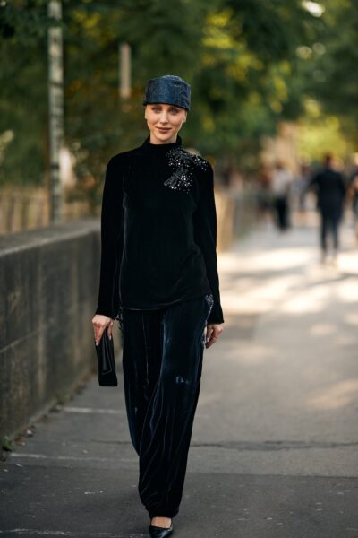 Zdjęcie przedstawia kobietę w ciemnej stylizacji z welurowego materiału, uzupełnionej czapką i minimalistyczną torebką. Scena rozgrywa się na miejskim chodniku, a rozmyte tło z przechodniami podkreśla naturalny, codzienny kontekst stylizacji. Kobieta nosi modne welurowe spodnie na zimę 2025. 