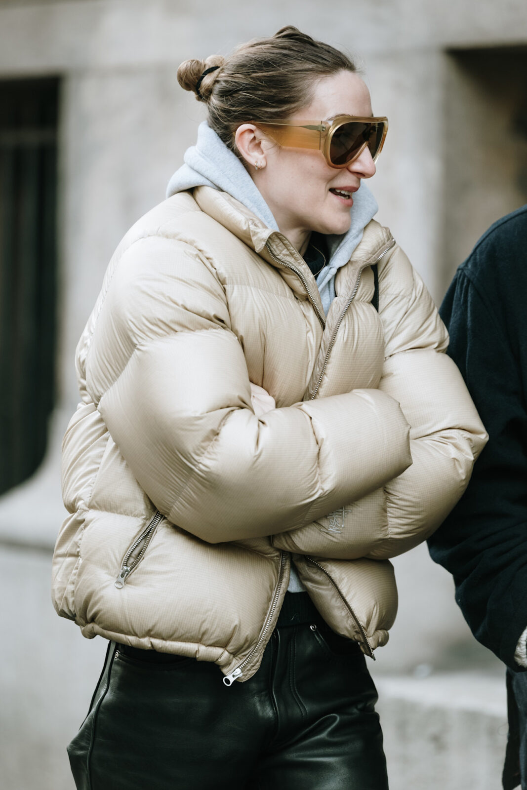 Zima 2026 trendy: Kobieta w czarnym, lakierowanym płaszczu o fakturze skóry krokodyla i dużej czapce z futrzanym wykończeniem, pozująca przy ścianie z kamienia. Jej dłonie spoczywają w kieszeniach, a fale blond włosów spływają spod nakrycia głowy – stylizacja w duchu luksusowego glamour inspirowanego rosyjską zimą i estetyką haute couture.