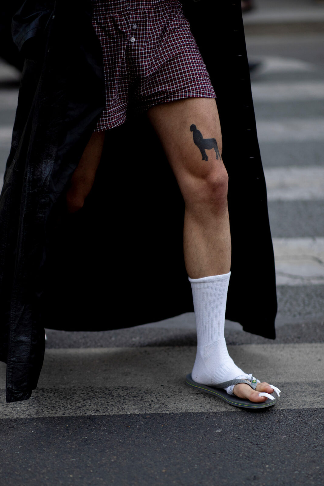 Havaianas trend: Moda uliczna podczas Paris Fashion Week – mężczyzna w białych skarpetkach i japonkach, ubrany w bordowe bokserki w kratę i długi czarny płaszcz. Na udzie widoczny tatuaż sylwetki psa, całość uchwycona w ruchu, w klimacie nonszalanckiego streetwearu.