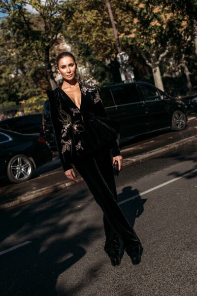 Zdjęcie przedstawia kobietę stojącą na ulicy, ubraną w czarną, welurową stylizację z głębokim dekoltem i ozdobnymi aplikacjami na ramieniu. W tle widoczne są zaparkowane samochody i drzewa, co nadaje całości charakter ulicznego street style’u inspirowanego pokazami mody.