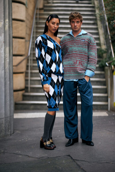 Para w stylizacjach retro z modowym zacięciem – kobieta w dzianinowym zestawie w niebiesko-czarno-białą kratę argyle, z ołówkową spódnicą i długimi podkolanówkami, oraz mężczyzna w luźnym, kolorowym swetrze z motywem vintage i jedwabistych, niebieskich spodniach. Zdjęcie wykonane na tle schodów w miejskim pejzażu, w duchu street style z Mediolanu lub Paryża.