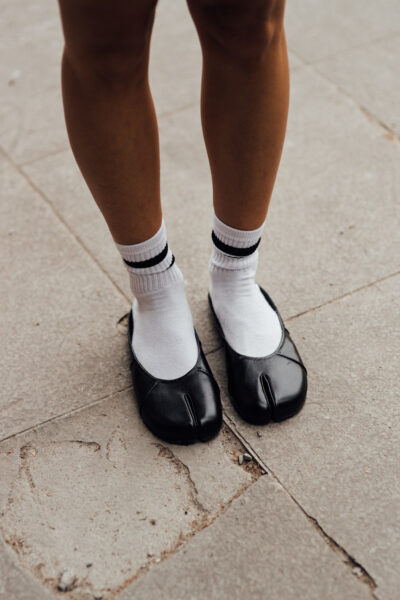 Zbliżenie na stopy w czarnych balerinach typu tabi, noszonych z białymi skarpetkami ze sportowymi paskami; minimalistyczny streetwearowy detal.