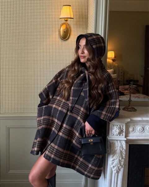 Elegancka kobieta w ciepłej, wełnianej pelerynie/ponczo w kratę (tartan) w odcieniach brązu, granatu i beżu, z kapturem lub kołnierzem szalowym. Stylizację dopełniają długie, czarne kozaki oraz mała, czarna torebka ze złotymi okuciami (quiet luxury). Tło stanowi klasyczne wnętrze z boazerią w kolorze szałwiowej zieleni oraz tapetą w drobną kratkę, oświetlone przez stylowy kinkiet.