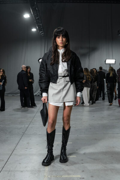 Kobieta w stylizacji streetwearowej na evencie modowym SHEIN w Mediolanie – uczestniczka w czarnej bomberce, białej koszuli i krótkiej spódnicy z ekoskóry, trzymająca kopertówkę i nosząca masywne czarne kozaki. W tle uczestnicy wydarzenia i industrialna przestrzeń hali pokazowej.