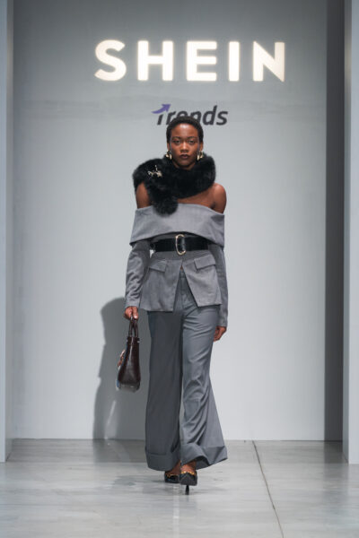 Modelka w stylizacji retro-chic na wybiegu SHEIN – look inspirowany modą lat 70.: zielony garnitur z szerokimi spodniami, biała koszula, fioletowy pasek i płaszcz w kolorze indygo. Dopełnieniem są kapelusz w kolorze khaki, duże okulary i mała torebka typu clutch.