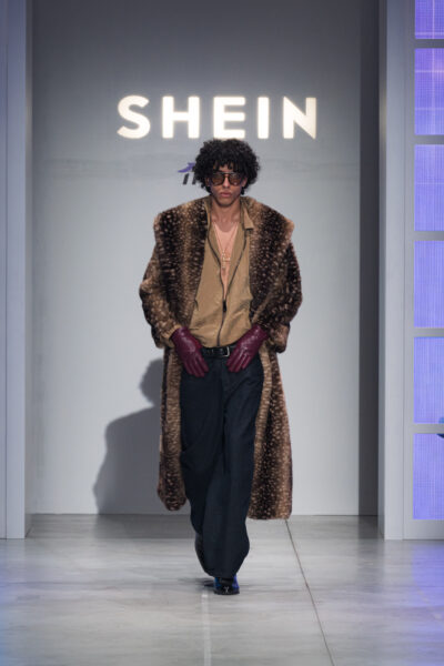 Modelka w szarym garniturze z odkrytymi ramionami podczas pokazu SHEIN – elegancka stylizacja z szerokim pasem, futrzanym szalem i torebką w kolorze burgundu. Mocny makijaż z ciemnymi ustami i złote kolczyki dodają całości luksusowego charakteru.