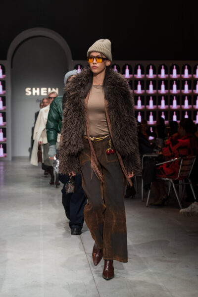 Model w płaszczu ze sztucznego futra na pokazie SHEIN – mężczyzna w długim brązowym futrze z białym wzorem, złotym naszyjnikiem i okularach przeciwsłonecznych. Stylizacja w klimacie retro z rozpiętą koszulą i bordowymi rękawiczkami.