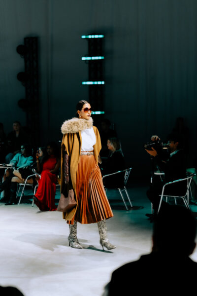 Modelka na wybiegu podczas pokazu SHEIN w Mediolanie – stylizacja w odcieniach miodowego brązu i bieli, z plisowaną spódnicą, futrzanym kołnierzem i wysokimi butami w zwierzęcy wzór. Kobieta w dużych okularach przeciwsłonecznych i eleganckim płaszczu, idąca po wybiegu wśród publiczności.