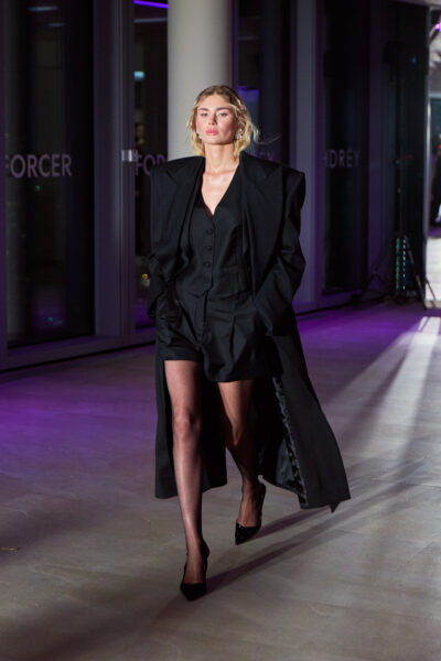 Stylizacja runway HDRÈY x FORCER w czerni – modelka w oversize’owej marynarce z poszerzanymi ramionami, klasycznych szortach garniturowych i czarnym topie na guziki. Look wykończony minimalistycznymi szpilkami.
