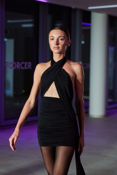 Czarna mini sukienka bandeau z marszczeniami i krzyżowanym halterem na szyi, prezentowana podczas pokazu HDRÈY x FORCER. Kreacja odsłania brzuch i ma mocno dopasowaną sylwetkę.