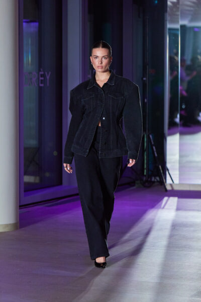 Oversize’owa stylizacja denimowa w czerni – szeroka kurtka jeansowa o podkreślonej linii ramion i proste spodnie, zestawiony ze szpilkami. Ujęcie z wybiegu HDRÈY x FORCER w futurystycznej oprawie świetlnej.