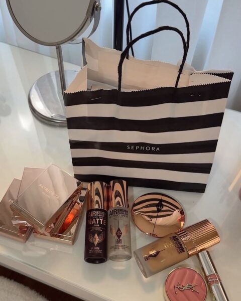 Zakupy kosmetyczne z Sephora ułożone na białej toaletce przed lustrem. Centralnym punktem jest duża, czarno-biała torba Sephora. Przed torbą widoczne są produkty w eleganckich, błyszczących opakowaniach w kolorze różowego złota i miedzi, w tym: trzy palety cieni lub róży Patrick Ta Beauty, dwie buteleczki podkładu Charlotte Tilbury Airbrush Flawless Matte oraz Airbrush Flawless Setting Spray, okrągły puder w różowym złocie (prawdopodobnie Charlotte Tilbury), podkład Charlotte Tilbury Airbrush Flawless Foundation oraz róż YSL i szminka.