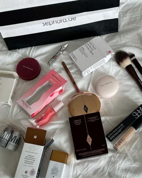 Widok z góry na zakupy kosmetyczne z Sephora, rozłożone na białej pościeli, z widoczną w tle górną częścią czarno-białej torby Sephora. Wśród produktów znajdują się: dwa opakowania od Rare Beauty (róż w pudełku Soft Pinch Luminous Powder Blush i puder), tusz do ust Gisou Honey Infused Lip Oil, podkład Charlotte Tilbury Airbrush Flawless Finish, korektor Nars Radiant Creamy Concealer, produkty do pielęgnacji Drunk Elephant (w tym D-Bronzi Anti-Pollution Sunshine Drops) oraz pędzle do makijażu Hourglass.