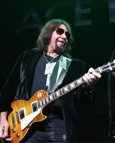 Gitarzysta Ace Frehley, dawny członek zespołu KISS, na scenie. Ma długie, ciemne włosy, okulary przeciwsłoneczne typu aviator i charakterystyczną koziej bródkę. Jest ubrany w aksamitną, czarną marynarkę, czarną koszulkę i spodnie. Gra na brązowo-żółtej gitarze typu Les Paul, trzymając ją na ozdobnym pasku. W tle widoczna jest perkusja.