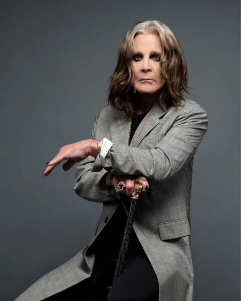 Portret muzyka Ozzy'ego Osbourne'a, siedzącego i opierającego się o elegancką laskę ze złotą rączką. Ma na sobie długi, szary płaszcz w kratę, czarne spodnie i czarny top. Spod rękawa płaszcza wystają białe, mankietowe koszuli. Muzyk ma pomalowane na czarno paznokcie, liczne pierścienie i charakterystyczną długą fryzurę z siwymi i brązowymi pasmami. Tło jest jednolicie szare.