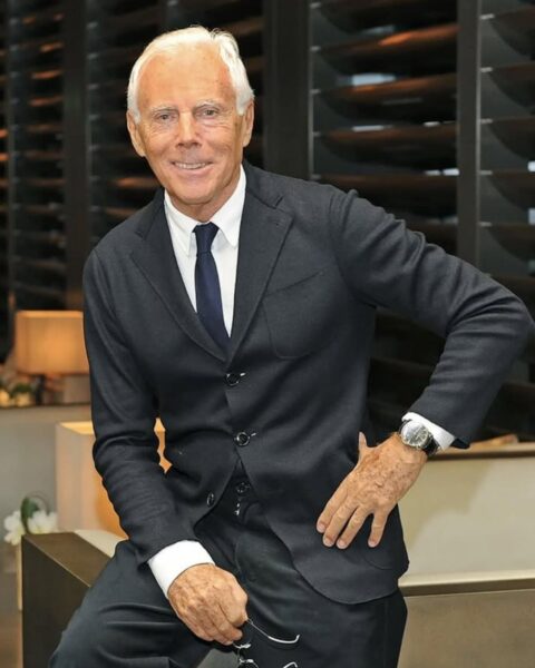 Portret włoskiego projektanta mody Giorgio Armaniego, ubranego w doskonale skrojony, ciemny garnitur (lub marynarkę) i białą koszulę z granatowym krawatem. Uśmiecha się, siedząc swobodnie i opierając jedną rękę na biodrze. Ma charakterystyczne, siwe włosy, a na nadgarstku widać elegancki zegarek. W ręce trzyma okulary. Tło jest nowoczesne i stonowane, z drewnianymi panelami i lampą.